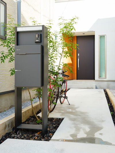 重厚なモダンテイストの玄関扉周りには木を貼り、宅配BOXを備えたポストにはシンボルツリーの緑。有機物と無機物を組み合わせたアプローチ